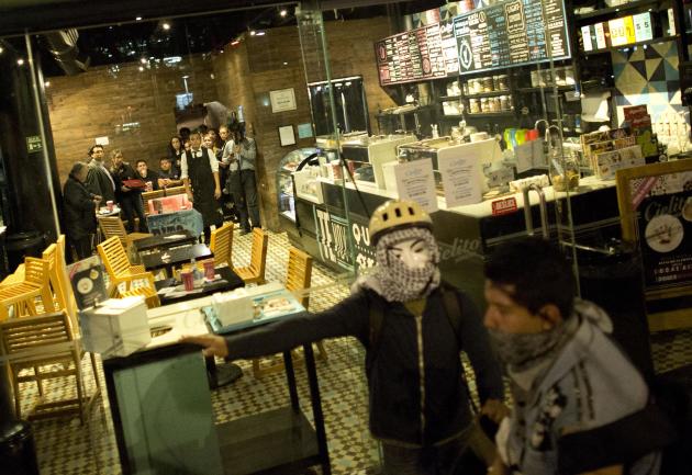 En esta imagen del 1 de diciembre de 2014, un manifestante anarquista dice a otros que no ataquen una cafetería, donde varias personas se reúnen al fondo, mientras los manifestantes rompen ventanas y 